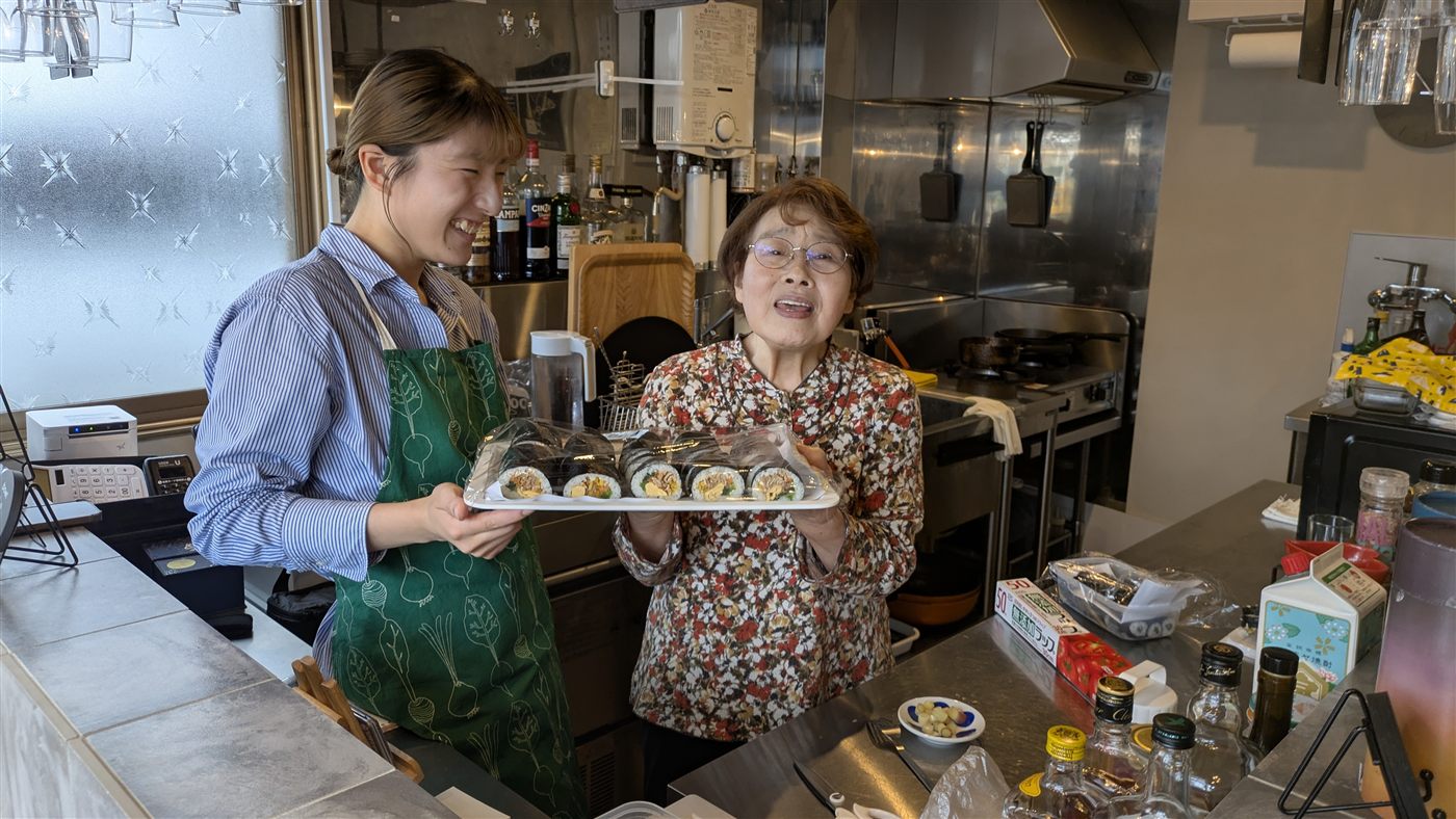 間借り居酒屋の厨房で、キンパの盛り合わせを持って笑うシニアの女性と学生